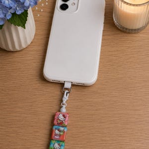 Puede incluir: Una funda de smartphone blanca con un colgante decorativo sujeto en la parte inferior. El colgante cuenta con tres cuentas cuadradas, cada una con un gráfico colorido de Hello Kitty, conectadas por un cierre de mosquetón blanco y una cuenta pequeña.