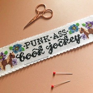 May include: White cross-stitch embroidery with a white lace border. The embroidery features a phrase "Punk-ass Hockey" in black text, two brown horses, and blue and purple flowers.