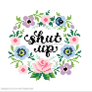 Puede incluir: Un patrón de punto de cruz con una corona de flores y el texto "Shut Up" en letras negras.