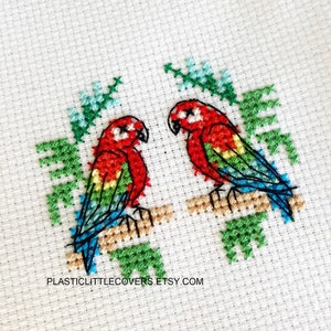 Puede incluir: Bordado de punto de cruz de dos loros rojos, verdes y azules posados ​​en ramas con hojas verdes. El texto "PLASTICLITTLECOVERS.ETSY.COM" está en la parte inferior de la imagen.
