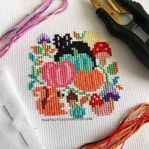 May include: Cross stitch pattern featuring a black cat, pumpkins, mushrooms, and a squirrel. The pattern is surrounded by green leaves and vines. The text "plasticlittlecovers.etsy.com" is at the bottom of the pattern.