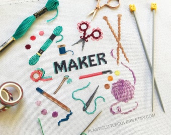 May include: Cross stitch pattern for a craft project featuring the word "MAKER" surrounded by various craft supplies, including scissors, yarn, needles, and tape. The pattern is printed on white fabric with a grid pattern.