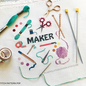 May include: Cross stitch pattern for a craft project featuring the word "MAKER" surrounded by various craft supplies, including scissors, yarn, needles, and tape. The pattern is printed on white fabric with a grid pattern.