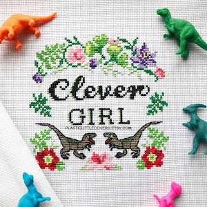 Könnte beinhalten: Kreuzstichmuster mit einem floralen Rand und dem Text "Clever Girl" in Schwarz. Zwei grüne Dinosaurierfiguren befinden sich auf dem weißen Stoff, sowie eine rosa und eine blaue Dinosaurierfigur.