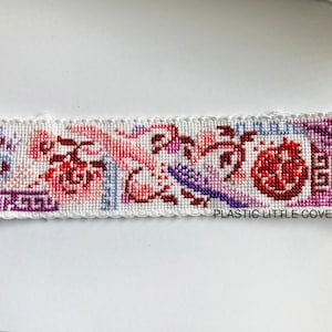 Op de afbeelding: Een witte kruissteek-bladwijzer met een bloemmotief met roze, rode en paarse bloemen, bladeren en een granaatappel. De bladwijzer heeft een witte gehaakte rand en de tekst "PLASTIC LITTLE COVERS" onderaan.