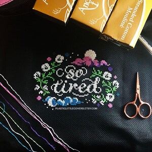 May include: A black fabric with a cross-stitch design that reads "So tired". The design is surrounded by a floral border. There are several spools of embroidery floss and a pair of small scissors on the fabric.