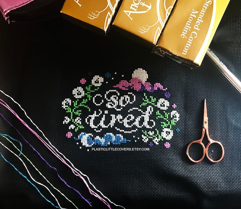 May include: A black fabric with a cross-stitch design that reads "So tired". The design is surrounded by a floral border. There are several spools of embroidery floss and a pair of small scissors on the fabric.