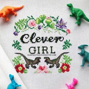 May include: Cross stitch pattern featuring a green, purple, and pink floral design with the text "Clever Girl" in black. The pattern is surrounded by green leaves and flowers.