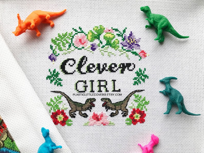 May include: Cross stitch pattern featuring a green, purple, and pink floral design with the text "Clever Girl" in black. The pattern is surrounded by green leaves and flowers.