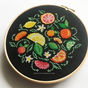 May include: A black embroidery hoop with a colorful cross-stitch design of various citrus fruits, flowers, and leaves. The design includes lemons, limes, oranges, grapefruits, and white flowers.