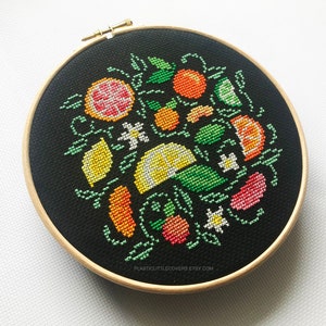 May include: A black embroidery hoop with a colorful cross-stitch design of lemons, limes, oranges, and other citrus fruits. The design is surrounded by green leaves and white flowers.