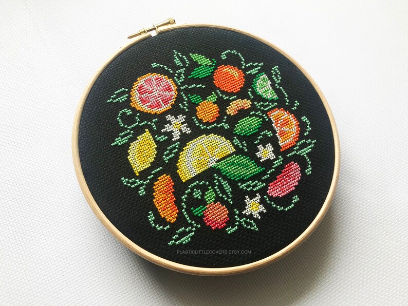 May include: A black embroidery hoop with a colorful cross-stitch design of lemons, limes, oranges, and other citrus fruits. The design is surrounded by green leaves and white flowers.