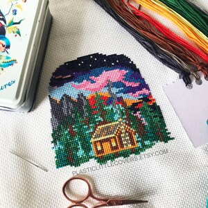 May include: A cross-stitch pattern of a cabin in the woods at night with a pink sky and stars. The pattern is on white Aida cloth and includes a needle, scissors, and floss.