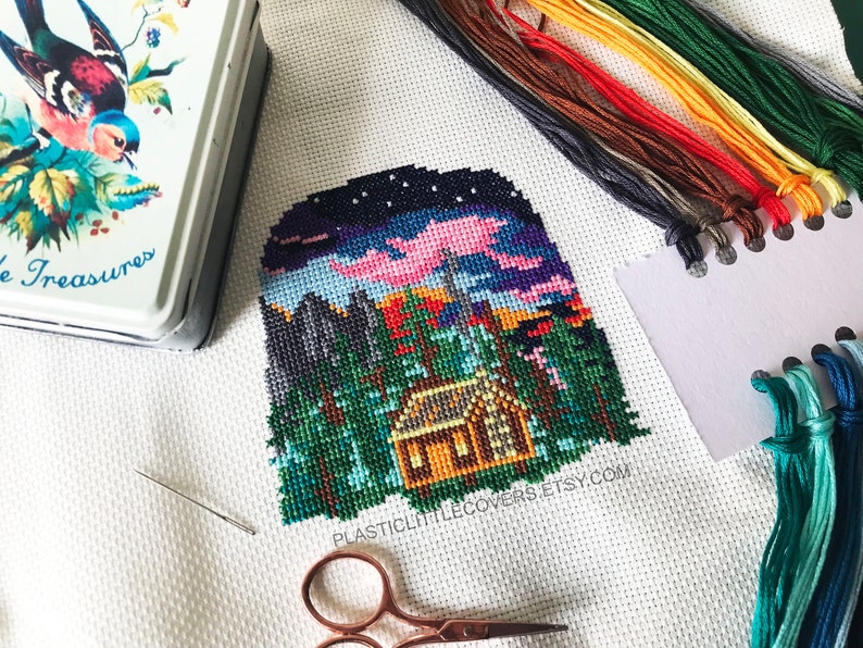 May include: A cross-stitch pattern of a cabin in the woods at night with a pink sky and stars. The pattern is on white Aida cloth and includes a needle, scissors, and floss.