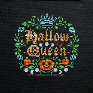 May include: Cross-stitch pattern featuring the text "Hallow Queen" in a gold color with a crown and pumpkins. The pattern is surrounded by green leaves and white moons.