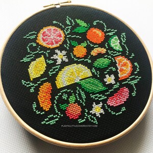 May include: A black embroidery hoop with a colorful cross-stitch design of lemons, limes, oranges, and grapefruit. The design is surrounded by green leaves and white flowers.