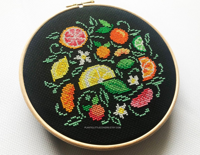 May include: A black embroidery hoop with a colorful cross-stitch design of lemons, limes, oranges, and grapefruit. The design is surrounded by green leaves and white flowers.
