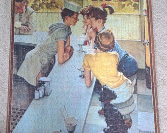 Könnte beinhalten: Ein rechteckiges Puzzle mit einer Illustration von Norman Rockwell für The Saturday Evening Post. Die Szene zeigt eine Sodatheke mit einem Kellner, zwei jungen Leuten und einem kleinen schwarzen Hund. Der Text lautet The Saturday Evening Post und Norman Rockwell.