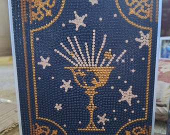 Könnte beinhalten: Ein rechteckiges Diamant-Malerei-Display mit dunkelblauem Hintergrund und einem goldenen Kelch in der Mitte. Das Design umfasst goldene Filigran-Ecken, weiße und goldene Sterne sowie weiße Strahlen, die vom Kelch ausgehen, alles in einem klaren Rahmen.