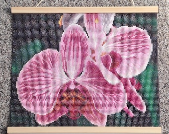 Könnte beinhalten: Ein quadratisches Diamond-Painting-Wandbild, das eine detaillierte rosa Orchideenblüte mit dunkelvioletten und gelben Akzenten zeigt. Das Kunstwerk ist zwischen zwei hellen Holzleisten oben und unten befestigt und hat eine dünne Kordel.