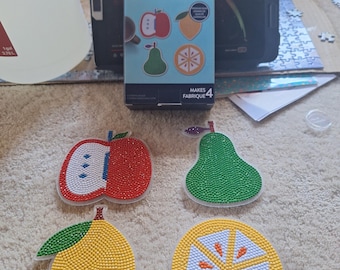 Könnte beinhalten: Ein Jonathan Adler x Michaels Diamond Art Untersetzer-Set mit vier fruchtförmigen Untersetzern: ein roter Apfel, eine grüne Birne, eine gelbe Zitrone und eine Zitrusfruchtscheibe. Die Schachtel zeigt den Text Jonathan Adler x Michaels Diamond Art Coaster Kit.