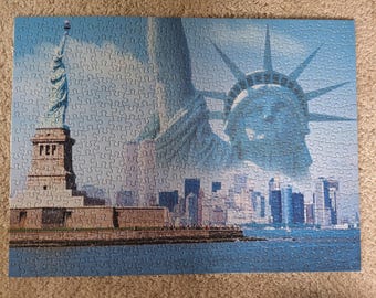 Könnte beinhalten: Ein rechteckiges Puzzle mit einer blau getönten Montage der Freiheitsstatue auf ihrem Sockel, einer großen Nahaufnahme des Kopfes der Statue mit ihrer Krone und der Skyline von New York City vor einem klaren Himmel und Wasser.