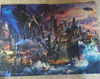 Könnte beinhalten: Ein rechteckiges Puzzle mit einer detaillierten Illustration von Gotham City bei Nacht. Batman fliegt über dem Hafen, während Aquaman aus einer großen Welle auftaucht. Die Szene zeigt einen Dampfzug, ein Frachtschiff und ein Luftschiff.