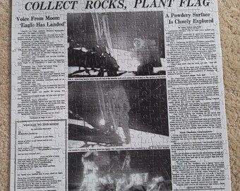 Könnte beinhalten: Ein Puzzle mit einer Reproduktion der Titelseite der New York Times vom 21. Juli 1969. Die Schlagzeile lautet MEN WALK ON MOON, ergänzt durch Unterüberschriften und Schwarz-Weiß-Fotos der Mondlandung, der Astronauten und der Mondlandefähre.