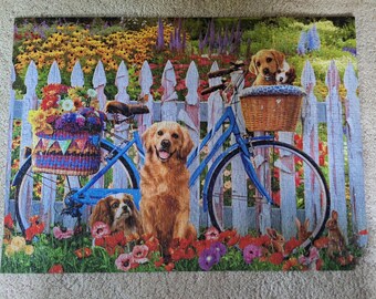 Könnte beinhalten: Ein Puzzle, das ein blaues Fahrrad mit einem Weidenkorb zeigt, in dem zwei Welpen sitzen, geparkt vor einem weißen Lattenzaun. Ein Golden Retriever sitzt im Vordergrund zwischen roten Mohnblumen, daneben ein kleiner Hund und Kaninchen.