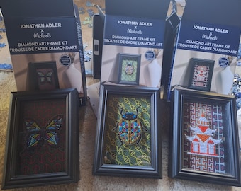 Könnte beinhalten: Drei Jonathan Adler x Michaels Diamond Art Rahmen-Sets mit schwarzen Rahmen. Die Designs zeigen einen bunten Schmetterling, einen mehrfarbigen Käfer und eine weiß-orange Pagode, jeweils vor gemusterten Hintergründen in schwarzen Rahmen.