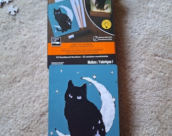 Könnte beinhalten: Ein Artist's Loft Malen-nach-Zahlen Acryl-Set mit einem schwarzen Katzenmotiv auf einer Mondsichel. Die Verpackung zeigt den Inhalt mit Farben, Pinseln und einer Leuchtvorrichtung. Eine fertig bemalte Platte steht auf einem Holzsockel.