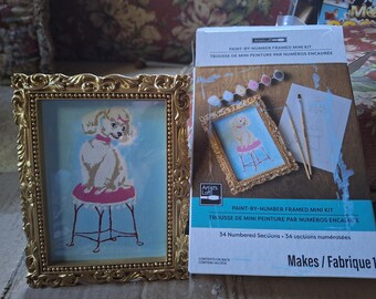 Könnte beinhalten: Ein Artist's Loft Malen-nach-Zahlen gerahmtes Mini-Set mit einem Pudel auf einem rosa Hocker. Das Set enthält einen goldfarbenen, verzierten Rahmen, zwei Pinsel und Farbtöpfe. Die Schachtel zeigt den Text Paint-By-Number Framed Mini Kit und 34 Numbered Sections.