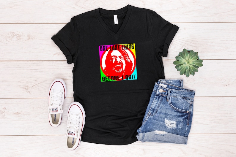 Pode incluir: Camiseta preta de gola em V com um gr&aacute;fico colorido de um rosto e o texto "One Last Thing Before I Quit".