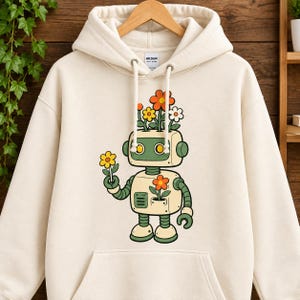 May include: A cream-coloured hooded sweatshirt featuring a graphic of a light-coloured robot with green accents. The robot has orange, yellow, and white flowers growing from its head and body, and it is holding a single yellow flower in its hand.