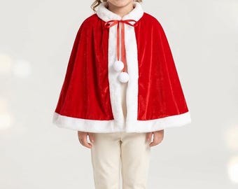 May include: A bright red velvet cape featuring a thick white faux fur trim along the collar and hem. The cape includes a red ribbon tie at the neck adorned with two white pom-poms. The garment is worn over a cream-colored long-sleeve top and pants.