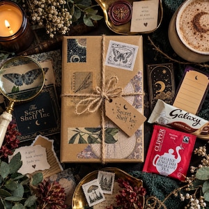 May include: A rectangular gift box wrapped in brown paper with twine, featuring a tag labeled The Secret Shelf Inc. The box is surrounded by a magnifying glass, a candle, a mug of coffee, a Galaxy chocolate bar, and a Clipper organic tea bag.