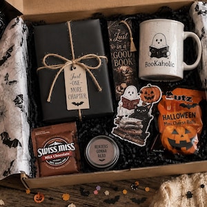 Puede incluir: Una caja de regalo con un libro envuelto en negro con una etiqueta Just One More Chapter, una taza blanca con un fantasma leyendo y el texto Bookaholic, un marcapáginas, una pegatina de fantasma y gato, cacao Swiss Miss y bolas de queso Utz.