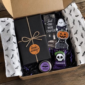 May include: A cardboard gift box lined with white tissue paper featuring black bats, containing a black wrapped gift tied with twine and a round orange sticker reading SPOOKY STORIES, a bookmark reading JUST ONE MORE CHAPTER, stickers, and a treat.