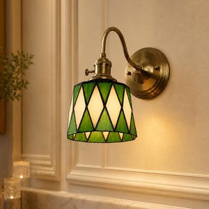 May include: A wall-mounted lamp featuring a brass-toned metal fixture with a curved arm and a circular base. The lampshade is constructed from stained glass panels arranged in a geometric diamond pattern, alternating between green and white colors.
