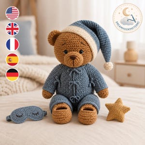May include: A brown crocheted teddy bear wearing a blue pajama set with tied closures and a matching blue nightcap with a cream-colored brim and pom-pom. Beside the bear sits a blue crocheted sleep mask with embroidered eyes and a small yellow star.