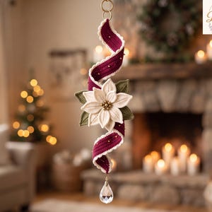 Op de afbeelding: Een hangend gehaakt ornament met een centrale witte kerststerbloem met groene bladeren, bevestigd aan een spiraalvormig donkerrood lint met witte geschulpte randen en kleine gouden kralen, afgewerkt met een heldere druppelvormige kristallen hanger.