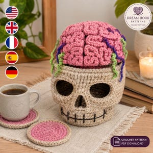 Könnte beinhalten: Ein beigefarbener, gehäkelter Totenkopf-Behälter mit einem abnehmbaren, rosafarbenen Gehirn-Deckel, der mit grünen und violetten Garnakzenten versehen ist. Der Totenkopf hat schwarze Stickereien an Mund und Augenhöhlen. Ein passender rosafarbener Untersetzer.