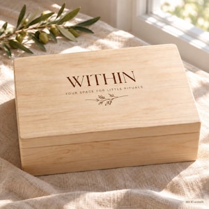 Könnte beinhalten: Eine rechteckige, helle Holzschatulle mit Klappdeckel. Auf der Oberseite befindet sich der dunkelbraune Schriftzug "WITHIN", gefolgt von "YOUR SPACE FOR LITTLE RITUALS" und einer kleinen, mittig platzierten Strichzeichnung eines Zweiges.