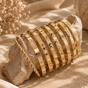 Peut inclure: Un ensemble de huit bracelets rigides en métal doré et un bracelet en chaîne torsadée dorée. Chaque bracelet rigide présente un motif gravé distinct, incluant des formes géométriques, des étoiles et des textures croisées, disposés en pile.