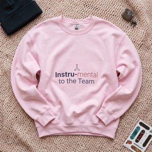 May include: A light pink crewneck sweatshirt featuring the text "Instru-mental to the Team" in dark blue and red lettering, with a small graphic of surgical scissors positioned above the word "mental" on the center of the chest.