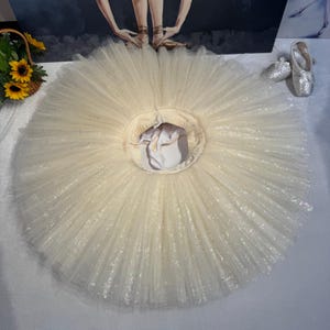 Puede incluir: Un tutú de ballet color crema compuesto por múltiples capas de tela de tul fino plisado con un acabado sutilmente brillante. La prenda presenta una silueta circular acampanada con una cintura elástica central y un forro de tela blanca integrado.