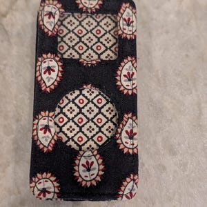 May include: A rectangular fabric case for a portable media player featuring a black background with a repeating pattern of red and white paisley motifs. The case includes a rectangular cutout for a screen and a circular cutout for a control wheel.