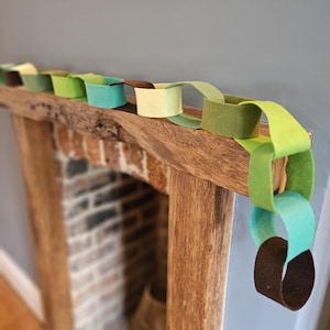 May include: A decorative garland made of interlocking felt loops in various shades of green, yellow, and brown. The chain consists of individual circular felt links connected together to form a continuous, flexible strand draped over a wooden beam.
