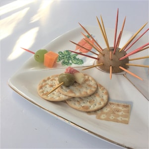 Puede incluir: Una bandeja de servir de cerámica blanca con cuatro secciones. La bandeja tiene un diseño floral y un borde dorado. La bandeja está llena de aperitivos, incluyendo galletas, aceitunas, queso y una patata con palillos de dientes insertados en ella.