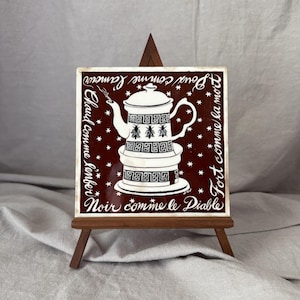May include: A square ceramic tile featuring a dark brown background with a white illustration of a coffee pot decorated with bees and geometric patterns. The tile displays the French text: Doux comme l'amour, Chaud comme l'enfer, Fort comme la mort, Noir comme le Diable.